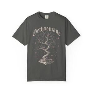 May include: Dark gray t-shirt featuring a stylized tree with pink blossoms and the word "Gethsemane" in a decorative font. The tree is set on a floating island. A casual, comfortable tee.