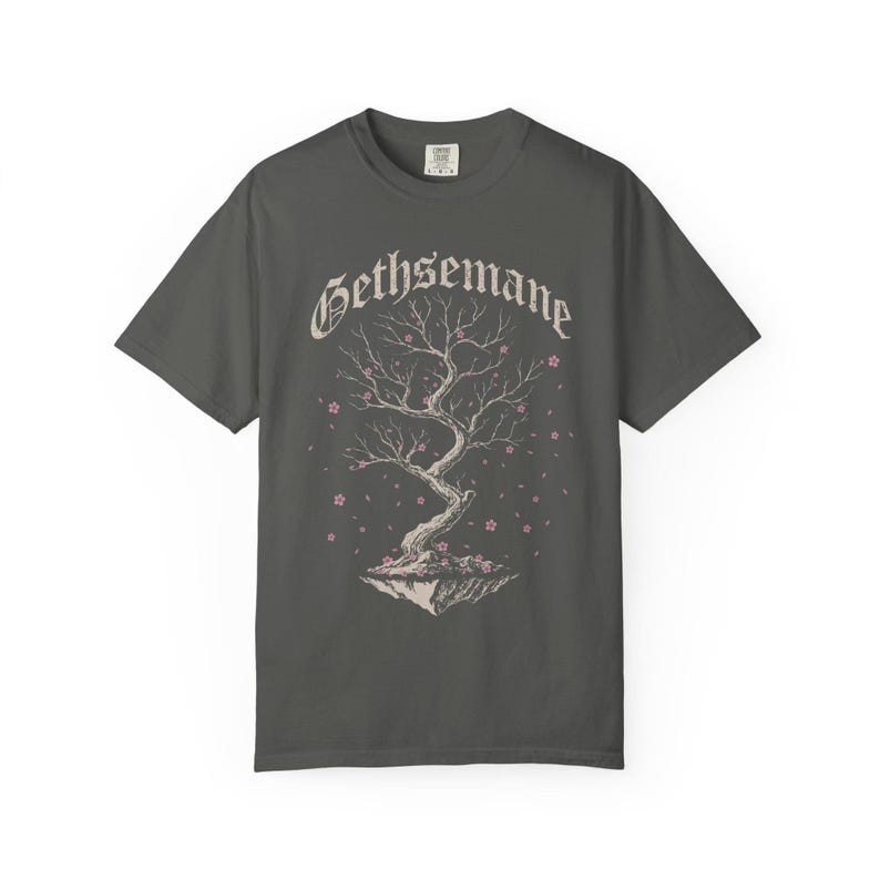 May include: Dark gray t-shirt featuring a stylized tree with pink blossoms and the word "Gethsemane" in a decorative font. The tree is set on a floating island. A casual, comfortable tee.