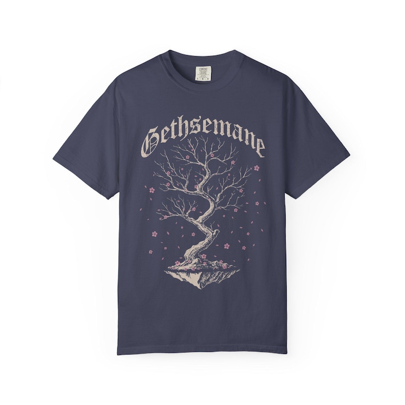 May include: A dark blue short-sleeve t-shirt featuring a graphic of a gnarled tree on a floating rock island with small pink blossoms. The word Gethsemane is printed in a white gothic-style font above the tree illustration on the front.