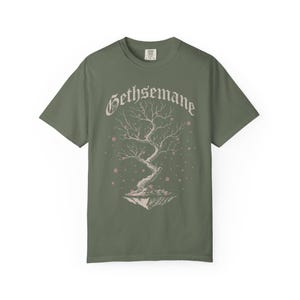 May include: Olive green t-shirt featuring the word "Gethsemane" in a stylized font above a detailed illustration of a tree with pink blossoms. The tree appears to be floating on a small island. The shirt is made of a soft material.