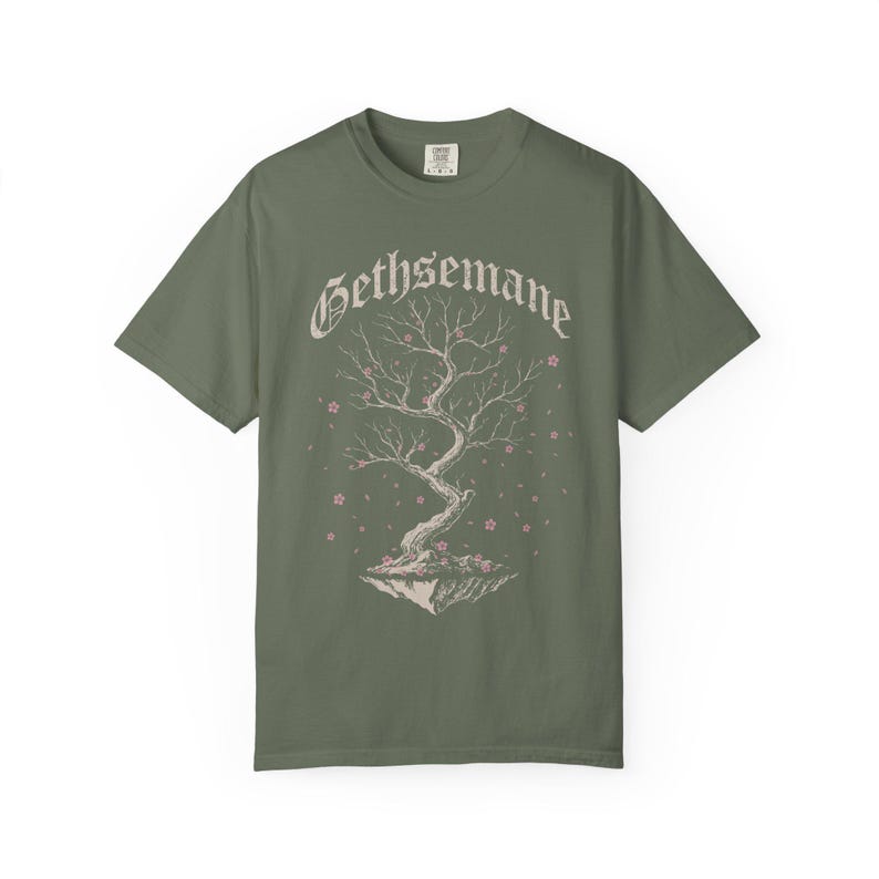 May include: Olive green t-shirt featuring the word "Gethsemane" in a stylized font above a detailed illustration of a tree with pink blossoms. The tree appears to be floating on a small island. The shirt is made of a soft material.
