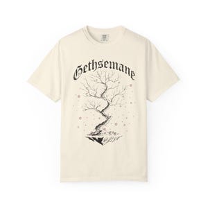 May include: Cream-colored t-shirt featuring a black illustration of a tree with the word "Gethsemane" above it. The tree is on a floating island with small star-shaped accents. The shirt has a classic crew neck and short sleeves.