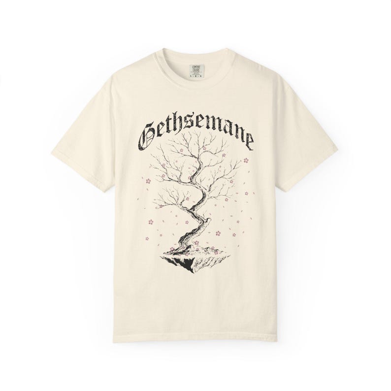 May include: Cream-colored t-shirt featuring a black illustration of a tree with the word "Gethsemane" above it. The tree is on a floating island with small star-shaped accents. The shirt has a classic crew neck and short sleeves.