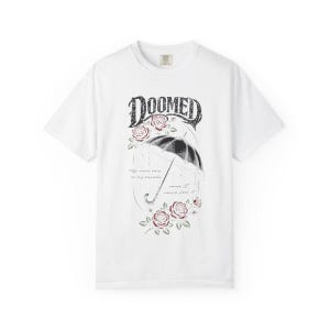 Pode incluir: Uma t-shirt branca de manga curta com uma impress&atilde;o gr&aacute;fica de um guarda-chuva preto, rosas vermelhas e o texto DOOMED no topo. Abaixo do guarda-chuva, o texto diz: So come rain on my parade, cause I wanna feel it. Design centralizado.