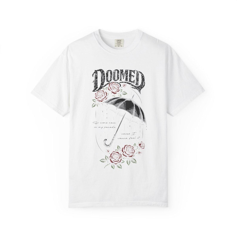 Pode incluir: Uma t-shirt branca de manga curta com uma impress&atilde;o gr&aacute;fica de um guarda-chuva preto, rosas vermelhas e o texto DOOMED no topo. Abaixo do guarda-chuva, o texto diz: So come rain on my parade, cause I wanna feel it. Design centralizado.