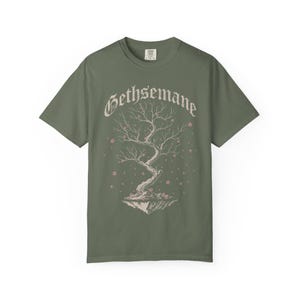 May include: An olive green short-sleeve t-shirt featuring a graphic of a bare tree on a floating island with small pink blossoms. The word Gethsemane is printed in a gothic-style font above the tree. The shirt has a crew neckline and short sleeves.