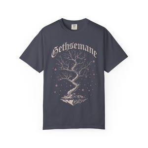 May include: A charcoal gray short-sleeve t-shirt featuring a graphic of a floating tree with bare branches and small pink blossoms. The word GETHSEMANE is printed in a gothic-style font above the tree illustration, which sits on a rocky base.