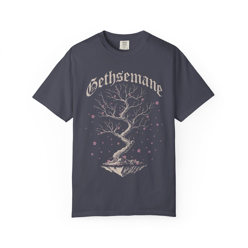 May include: A charcoal gray short-sleeve t-shirt featuring a graphic of a floating tree with bare branches and small pink blossoms. The word GETHSEMANE is printed in a gothic-style font above the tree illustration, which sits on a rocky base.