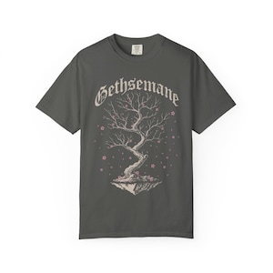 May include: A charcoal gray short-sleeve t-shirt featuring a graphic of a floating tree with twisted branches and small pink blossoms. The word Gethsemane is printed in a white gothic-style font above the tree illustration on the front center.