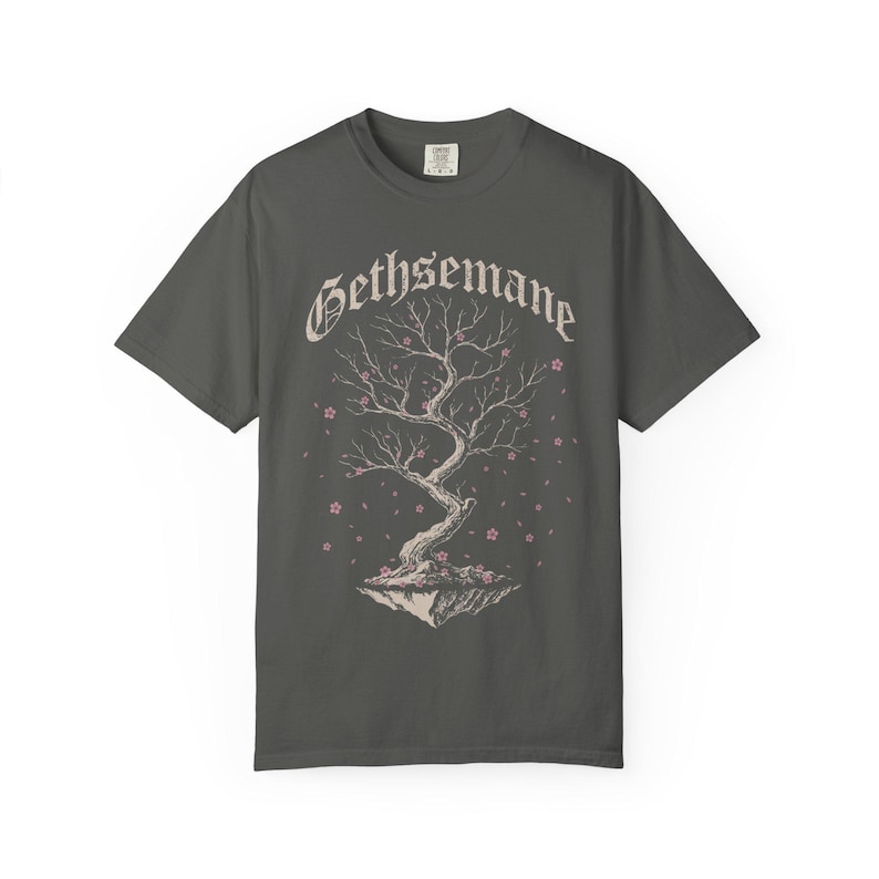 May include: A charcoal gray short-sleeve t-shirt featuring a graphic of a floating tree with twisted branches and small pink blossoms. The word Gethsemane is printed in a white gothic-style font above the tree illustration on the front center.