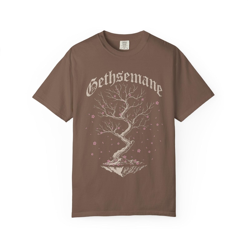 May include: A brown short-sleeve t-shirt featuring a graphic of a floating tree with bare branches and small pink blossoms. The word Gethsemane is printed in a white gothic-style font above the tree illustration, which sits on a small rock.