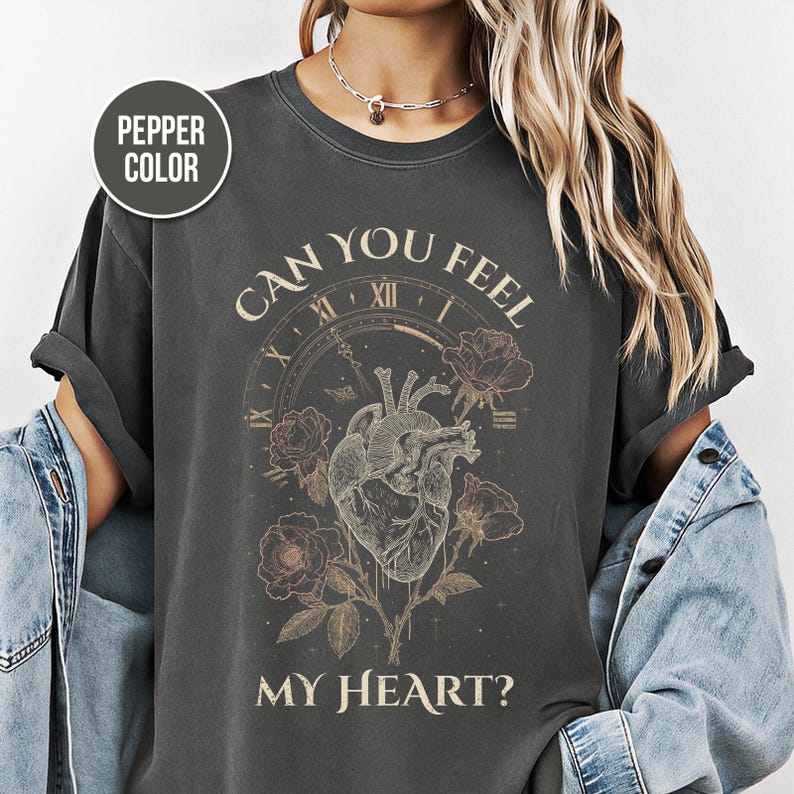 May include: Pepper-colored t-shirt with a graphic design featuring a human heart, roses, and the text "CAN YOU FEEL MY HEART?" The design includes a clock face. The shirt is a dark gray color.
