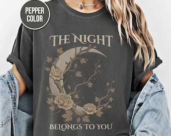 May include: A pepper-colored t-shirt featuring a graphic of a crescent moon entwined with bare branches and roses. The text reads THE NIGHT BELONGS TO YOU in a serif font. The design is rendered in muted earth tones against the dark gray fabric.