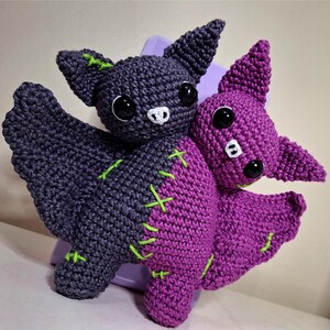 Pode incluir: Dois morcegos de crochê feitos à mão em tons de roxo e cinza. Os morcegos têm olhos grandes e pretos, narizes brancos e são costurados com linha verde neon. As asas estão abertas e os morcegos parecem estar descansando em uma superfície roxa.