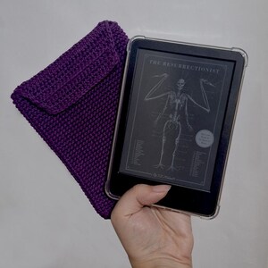 Pode incluir: Uma capa de leitor de e-books de crochê roxo escuro ao lado de um leitor de e-books exibindo o título "The Resurrectionist" e uma ilustração esquelética. O leitor de e-books está em uma capa protetora transparente.
