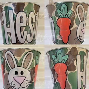 May include: Four metal buckets with a camouflage pattern, each with a different design. One bucket has the name "Hest" written on it, another has a bunny rabbit face, another has a carrot, and the last one has the letter "H" on it.