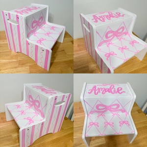 May include: A white two-step stool featuring pink painted details. The top step displays the name Amalie in pink script. The sides have vertical pink stripes, while the steps are decorated with a pink lattice pattern featuring small bows.