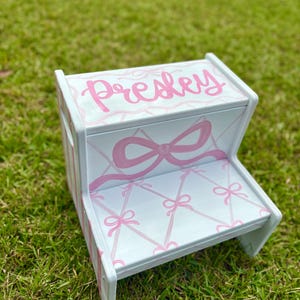 May include: A two-step wooden stool painted white with pink accents. The top step is inscribed with the name "Presley" in pink script. The steps and sides feature a bow and lattice design in pink.