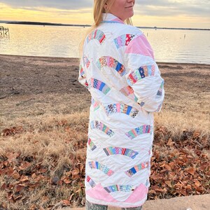 May include: A long, quilted coat featuring a patchwork design with white, pink, and various colorful fabric pieces. The coat has a unique, handcrafted appearance, with a distinct pattern and a comfortable, flowing silhouette.