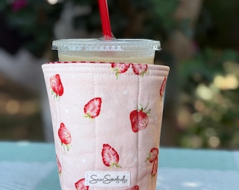 May include: A pink quilted drink sleeve adorned with red strawberries and white polka dots. A clear plastic cup with a lid and a red straw is inside the sleeve. The sleeve has a small white tag that reads "Sew Soulful".
