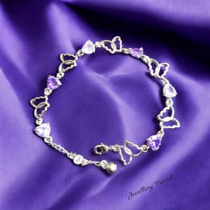 Può includere: Un braccialetto d'argento con maglie a forma di farfalla e pietre preziose viola e trasparenti a forma di cuore. Il braccialetto è esposto su uno sfondo di raso viola. La chiusura è aperta.