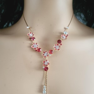 Può includere: Una collana d'oro con una catena delicata e accenti di fiori rossi e bianchi. La collana ha un ciondolo pendente con cristalli trasparenti.