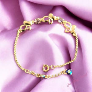 Puede incluir: Pulsera dorada con dijes en forma de mariposa y pequeñas gemas brillantes. El delicado diseño de la cadena incluye un cierre y una gema adicional al final. La pulsera se exhibe sobre una tela morada suave.