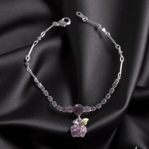 May include: A silver bracelet featuring a purple apple charm with a small green leaf. The bracelet includes small, round, purple beads and a larger purple gem. The background is a black satin fabric, creating a luxurious feel.