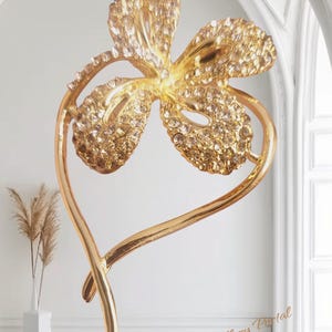 May include: A gold-toned decorative sculpture. The design features a flower with four crystal-studded petals, mounted on a curved stem. The text "Jewellery Portal" is visible in the lower right corner.