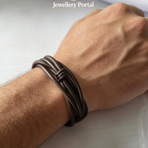 Puede incluir: Una pulsera de cuero marrón oscuro de múltiples hilos con un detalle de nudo central. La pulsera consta de varios cordones de cuero lisos y redondeados envueltos alrededor de la muñeca, creando una apariencia en capas con un diseño simple.