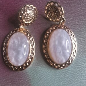 May include: A pair of gold-coloured earrings. Each earring has a textured, circular stud and an oval pendant with a pearlescent, white centre. The pendants are framed with a decorative gold border.