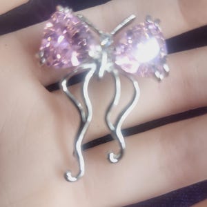 May include: A silver-coloured bow-shaped jewellery piece with a cluster of light purple gemstones. The bow has a delicate, open design with curved, dangling accents. The gemstones sparkle, catching the light.