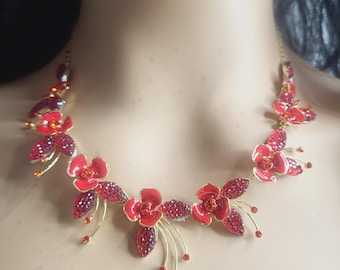 Könnte beinhalten: Eine goldfarbene Halskette mit einer Reihe von roten Emailleblumen und Blättern, die mit kleinen roten Kristallen verziert sind. Das florale Design ist symmetrisch entlang der Kette angeordnet, mit zarten goldfarbenen Drahtstielen an den Blüten.