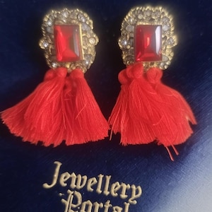 May include: A pair of ornate earrings with red rectangular gems and matching red tassels. The earrings have a gold-colored filigree setting with small, clear accent stones. The text "Jewellery Portal" is visible on the blue background.