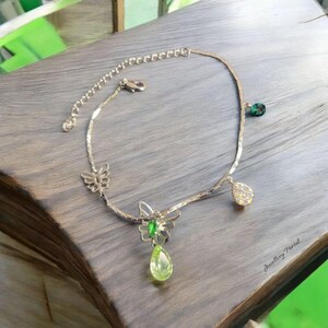 Puede incluir: Tobillera plateada con colgantes de mariposa y lágrima. La tobillera presenta una delicada cadena con un dije de mariposa y una gema verde en forma de lágrima. También hay gemas verdes y transparentes adicionales.