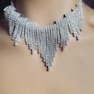 Pode incluir: Um colar choker prateado com um design em cascata de strass. O colar é feito de pequenos strass transparentes que são engastados em uma base de metal prateado.