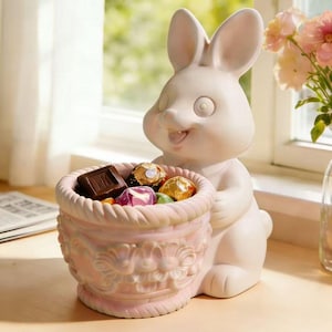 May include: A white ceramic bunny figurine holding a pink basket filled with assorted chocolates. The bunny has a smiling expression and large, round eyes. The basket has a decorative floral design.