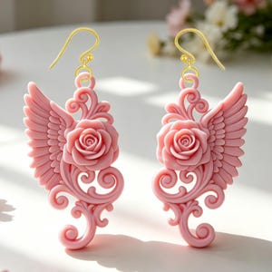 May include: A pair of pink earrings with a rose and wing design. The earrings have a gold-colored hook and feature a detailed rose in the center, flanked by wing-shaped accents. Intricate scrollwork adds to the decorative style.