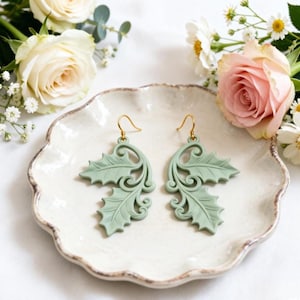 May include: A pair of sage green earrings with a holly leaf design. The earrings have a detailed, openwork pattern and gold-colored ear wires. The earrings are displayed on a white, scalloped-edge plate, surrounded by flowers.