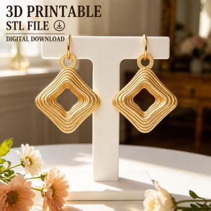 Può includere: Un paio di orecchini color oro con un design quadrato a righe concentriche e un foro centrale. Il testo 3D PRINTABLE STL FILE DIGITAL DOWNLOAD è visibile in alto. Gli orecchini sono esposti su un supporto bianco a forma di T.