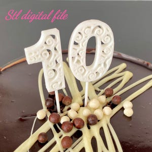 May include: A white cake topper shaped like the number 10 with an intricate swirling pattern, mounted on thin sticks. The topper is placed on a dark chocolate cake decorated with white chocolate drizzles and small brown and white candy spheres.