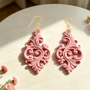 May include: A pair of pink drop earrings with gold-coloured ear wires. The earrings have a detailed, swirling design in a diamond shape. The earrings are displayed on a light-coloured surface, with soft shadows.