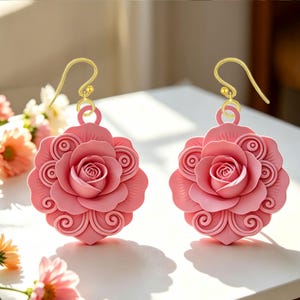 May include: A pair of pink rose-shaped earrings with gold-colored hooks. The earrings feature detailed petals and a central rose bud design. The earrings are displayed on a white surface, with soft lighting and shadows.