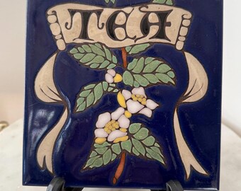 May include: A square, navy blue ceramic tile with the word "TEA" in black letters on a cream-colored banner. The tile features a floral design with white and yellow flowers, green leaves, and a brown stem. Cream-colored ribbons frame the banner and floral design.
