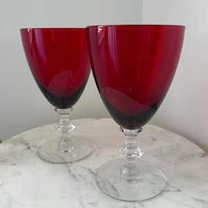 May include: Two ruby red wine glasses with clear stems and bases. The glasses have a classic design with a wide bowl and a slender stem. The glasses are made of glass and are likely used for serving wine.