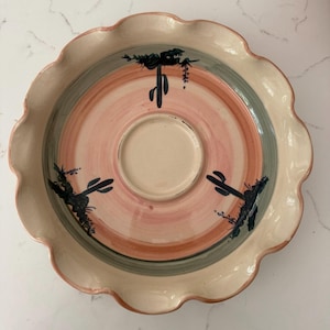 May include: A decorative ceramic serving dish with a scalloped edge. The dish features a central well and concentric rings of color in pink, green, and cream. Black cactus illustrations are painted around the center.