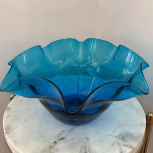 May include: A vibrant turquoise glass bowl with a ruffled rim. The bowl has a deep, rounded base and a flared, scalloped edge. The glass appears to be transparent, allowing light to pass through, and is likely used for decorative purposes.