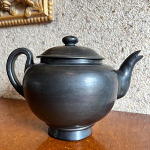 May include: A black teapot with a rounded body, a curved handle, and a spout. The lid has a small knob. The teapot is sitting on a wooden surface. The background is a textured wall and a gold-framed object.