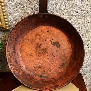 May include: A round copper skillet with a weathered, dark orange patina and visible surface markings. The pan features a sturdy metal handle attached to the rim with two rivets, showcasing a rustic, aged texture across the interior surface.
