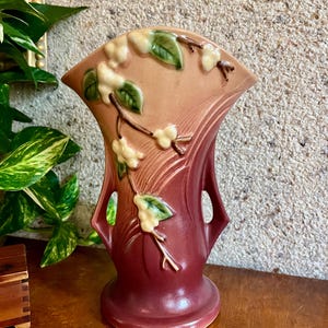 May include: A vintage ceramic vase in shades of burgundy, peach, and cream. The vase features a flared top with cut-out handles and is adorned with raised floral and leaf details. The design includes a rounded base and a textured finish, suitable for home decor.